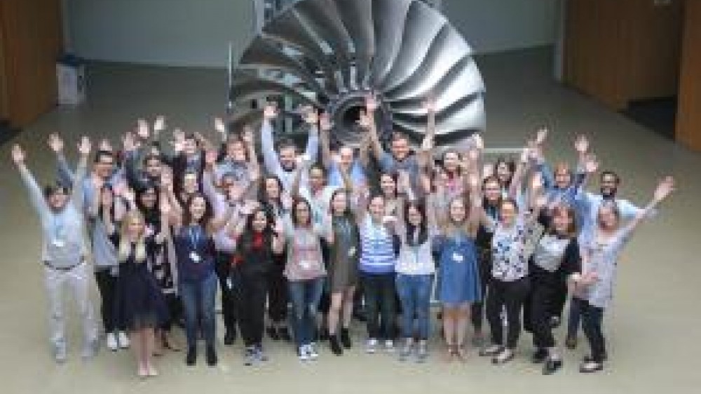 Students with there hands in the air standing in front of a large turbine 