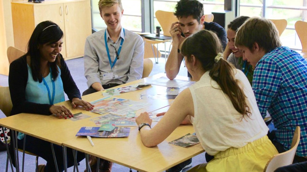 Students sitting around a table partaking in a public engagement exercises. The students were challenged to present their research using only pictures from magazines, and then using food