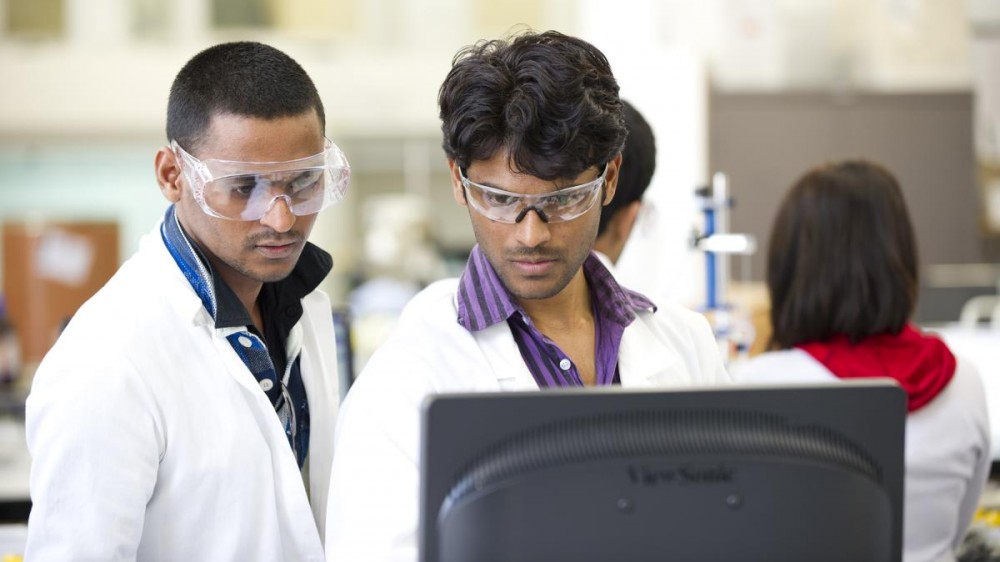 Two young male scientists in lab coats and goggles stand looking at a computer monitor.