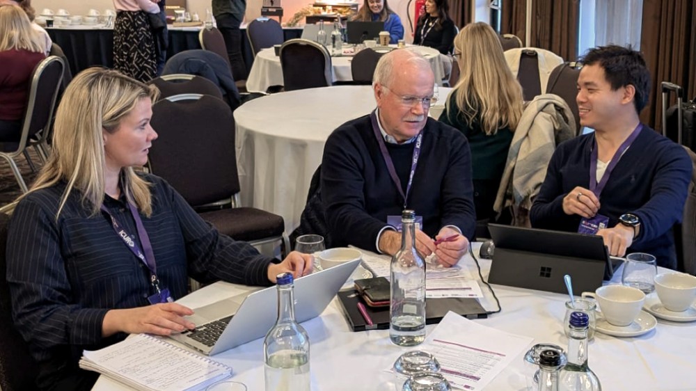 Three people at a table with papers and laptops in conversation