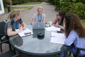 Three students sitting outside at a table. One of the students has a laptop and they are looking at her while in conversation