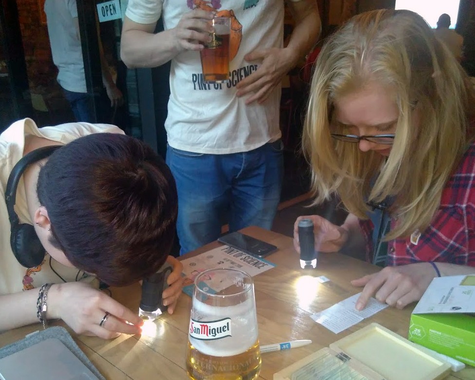 Two people sitting at a table looking down. They are using a device that has a torch to closely examine samples