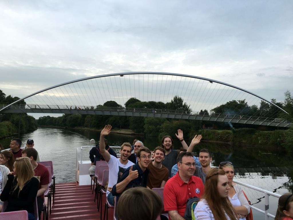 A group of students on a boat above deck. Some are waving at the camera. A bridge is in the background