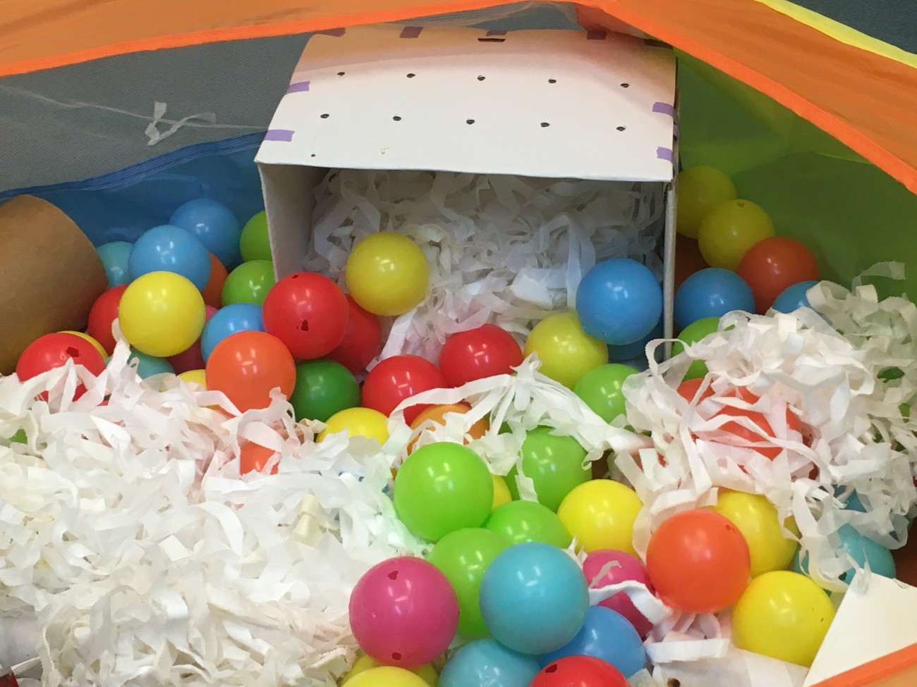 A perforated, cardboard transport carrier sits on its side in a playpen. The carrier is filled with strips of bedding material and surrounded by brightly coloured balls.