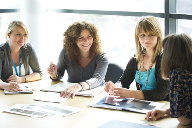 Four female researchers sit around a table discussing their research. You can see open books and paper on the table in front of them.