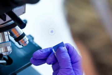 a photo showing a glass slide being held by a scientist, near to a microscope