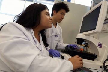 Deepali Pal using a microscope. A colleague is beside her looking at a screen