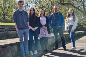 Five students standing by a low wall with tree's in the background